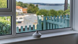 Ein Fenster mit einer weißen Vase auf dem Sims, Blick auf ein Gewässer und ein grüner Zaun im Hintergrund.