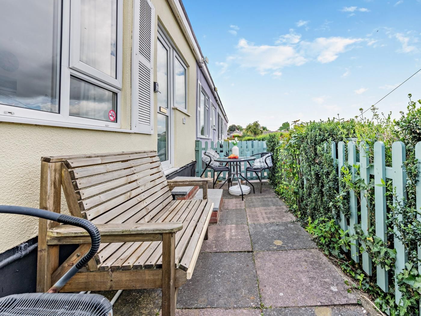 A wooden bench and a table with chairs in a garden area adjacent to a building with windows and shutters.