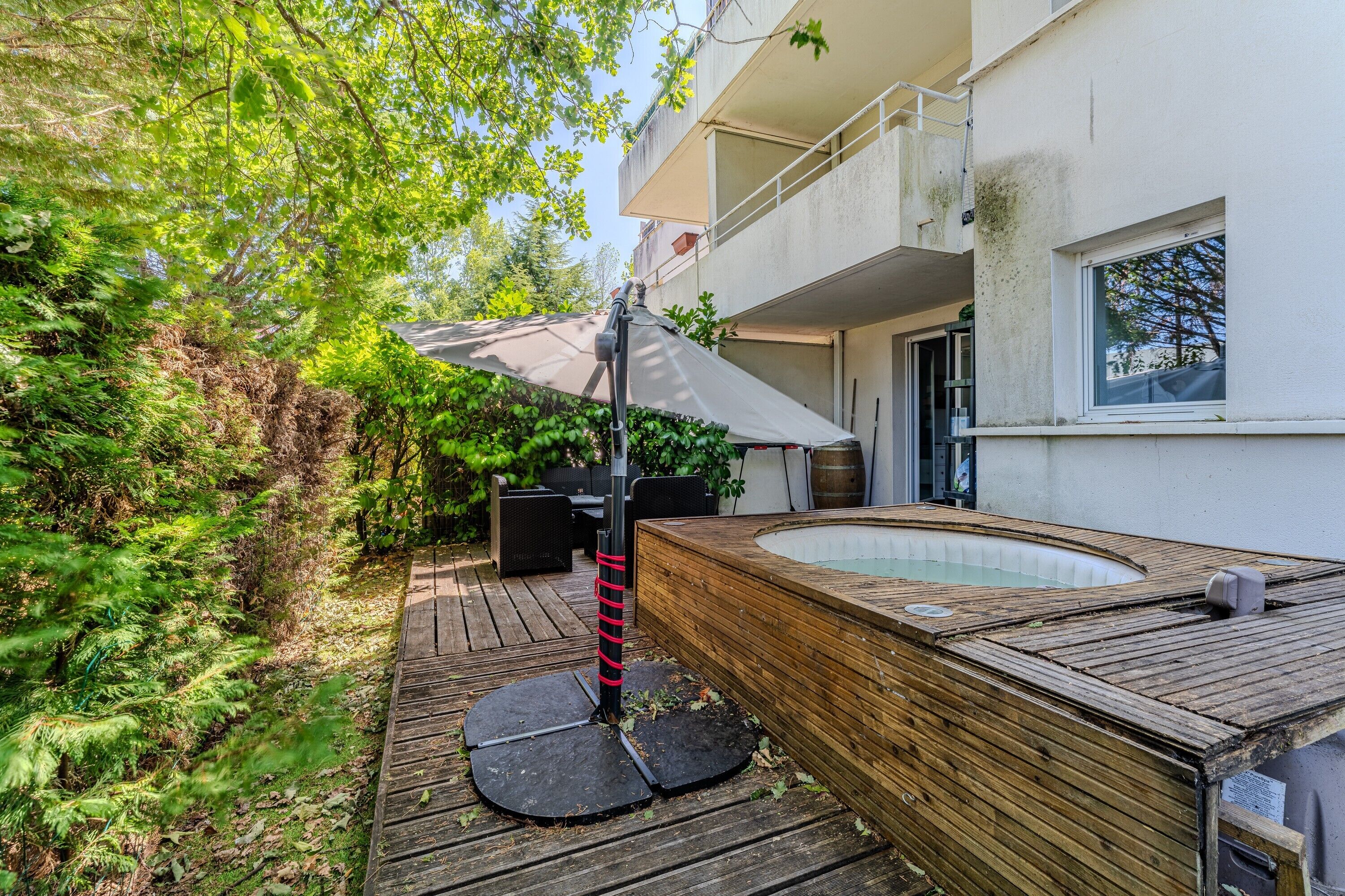 Eine Holzterrasse mit Whirlpool, umgeben von viel Grün und einem Gebäude mit Balkon.