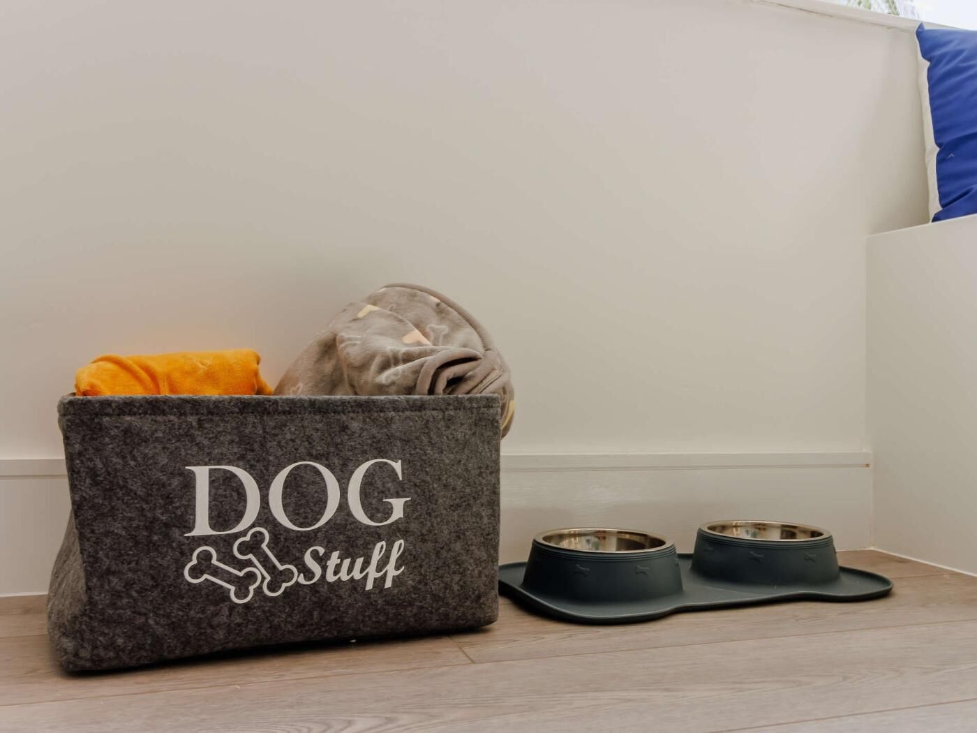 Un contenedor gris con la etiqueta 'Artículos para perros' que contiene una toalla amarilla y una manta gris, junto a dos cuencos negros para perros sobre un suelo de madera.