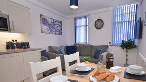 Ett modernt kök med vita skåp, en mikrovågsugn och en kaffebryggare. Det finns ett matbord med tallrikar, en skål med jordgubbar och croissanter. En grå soffa och en blå väggklocka syns också.