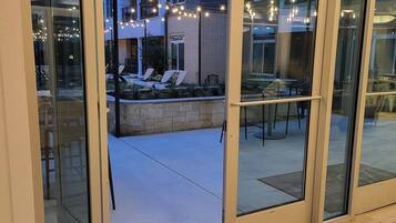 An entrance to a building with glass doors, a patio area with outdoor furniture, and a view of a multi-story building in the background.