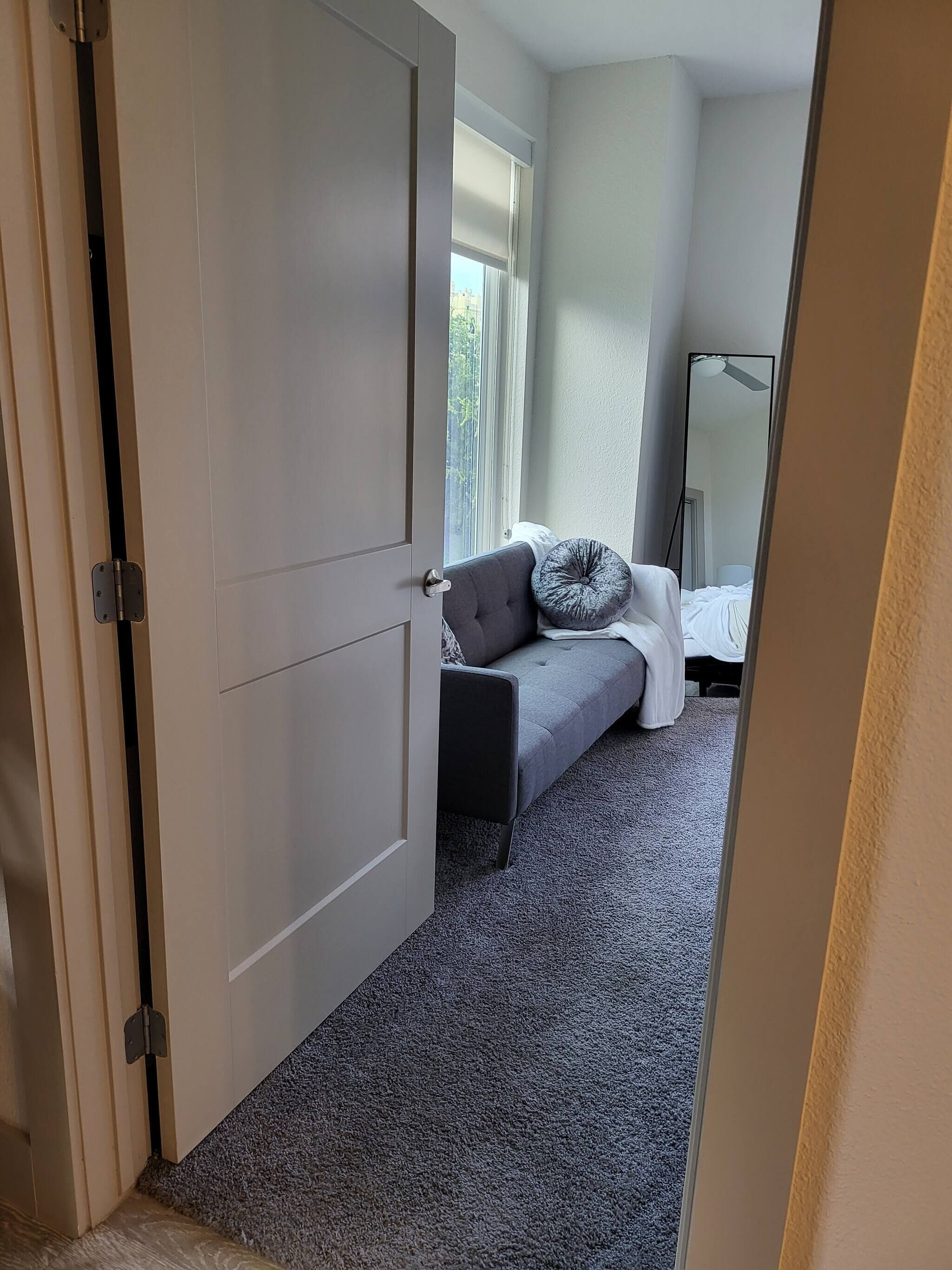 A bedroom with a bed, a grey sofa, a window, and a door.