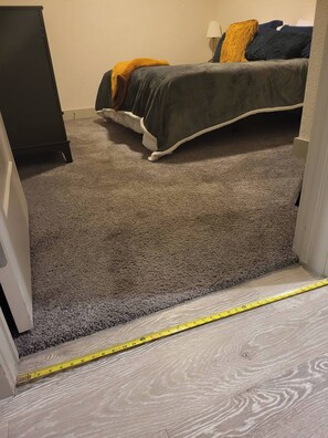 A carpeted room with a bed, a dresser, and a measuring tape on the floor.