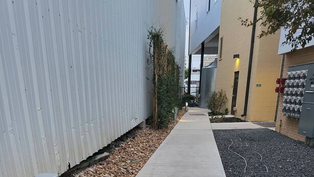 Um caminho de concreto ladeado por uma parede de metal e uma área ajardinada com pedriscos e grama.
