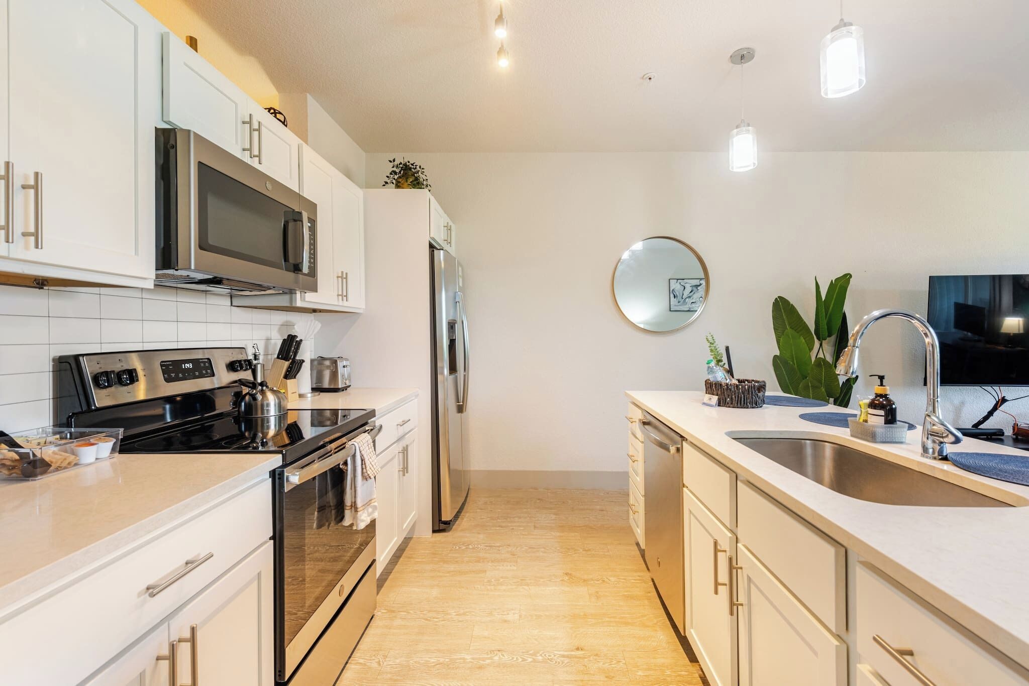 A modern kitchen with white cabinets, stainless steel appliances, a wooden floor, and a round mirror on the wall.