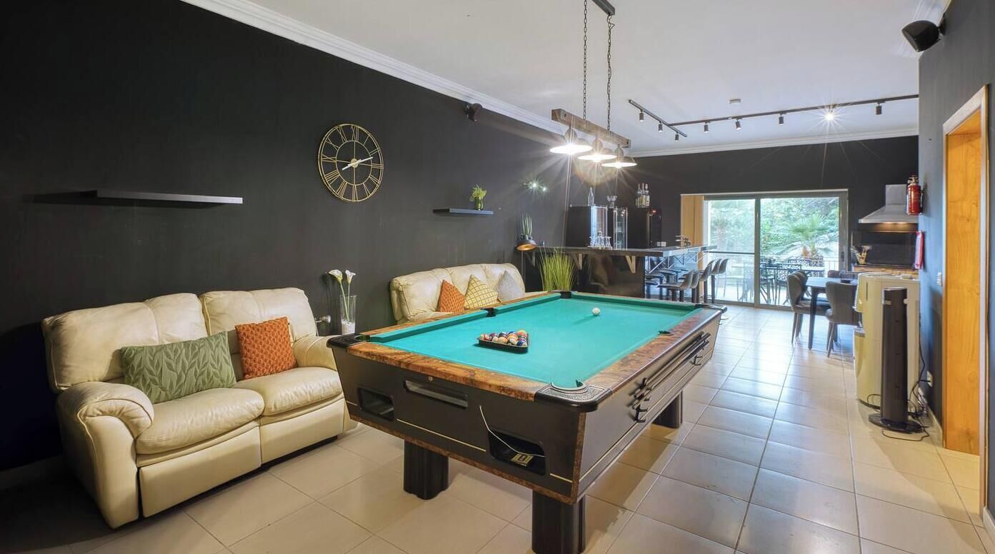 A billiards table with a pool cue and balls, a sofa with cushions, a clock on the wall, and a kitchen area in the background.
