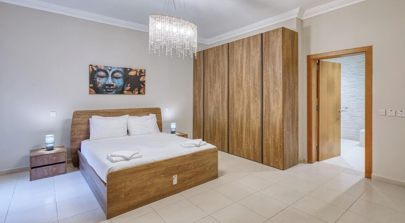 Un dormitorio con cama de madera, mesita de noche, una obra de arte en la pared, armario de madera y una puerta que da a otra habitación.
