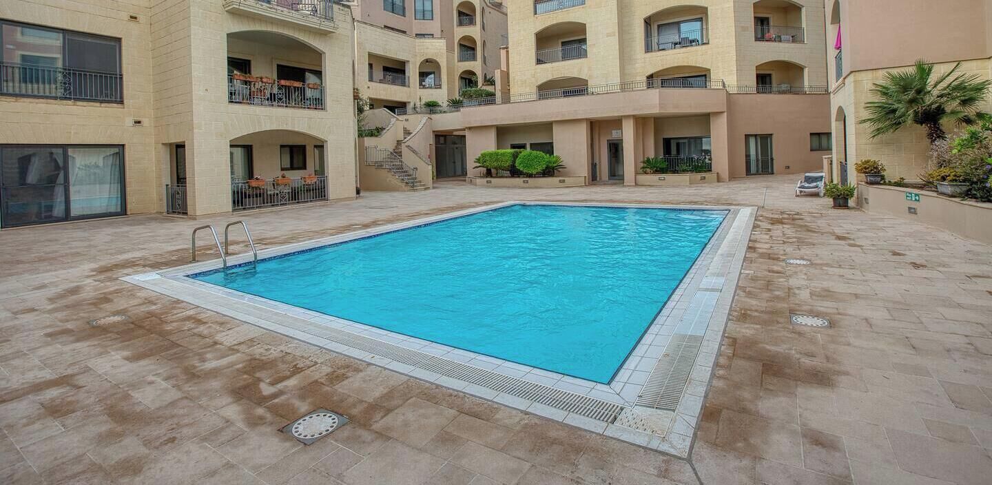 Una piscina con solárium empedrado, rodeada de edificios de varios pisos con balcones y mobiliario exterior.