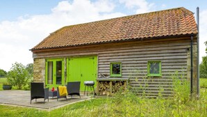 Una cabaña rústica de madera con techo de tejas, puerta verde y un pequeño patio con mobiliario exterior.