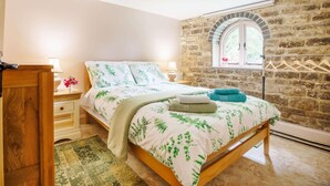 Un dormitorio con pared de piedra, cama de madera, mesita de noche, lámpara y ventana con vistas.