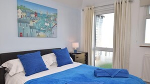 A hotel room with a bed, a painting of a coastal scene, a nightstand with a lamp, and a window with curtains. - 2 Bed in Pevensey (oc-55355) (Pevensey Bay)