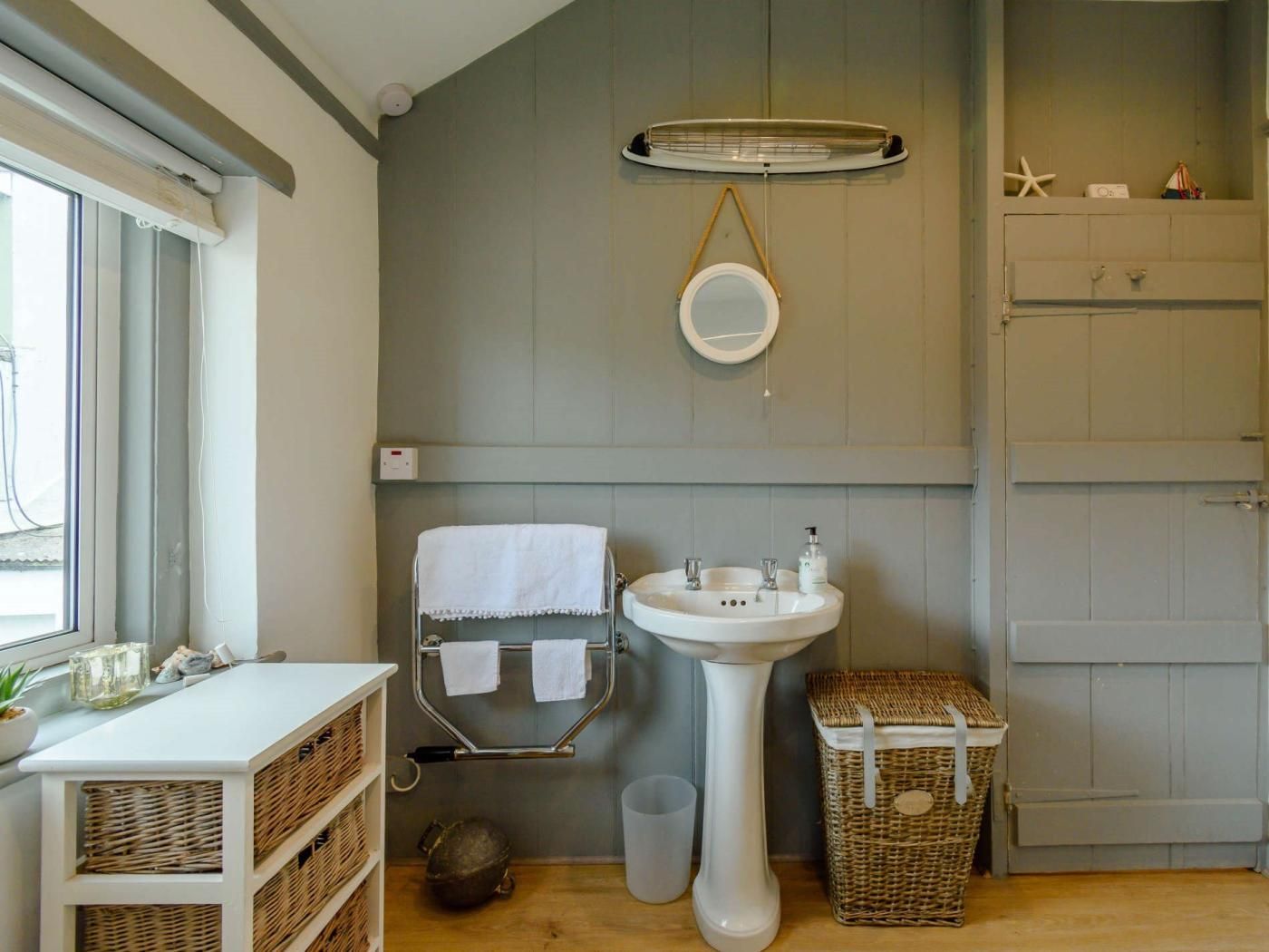 Une salle de bain avec un lavabo sur pied blanc, un porte-serviettes avec deux serviettes, un miroir rond, un panier en osier et une petite poubelle.
