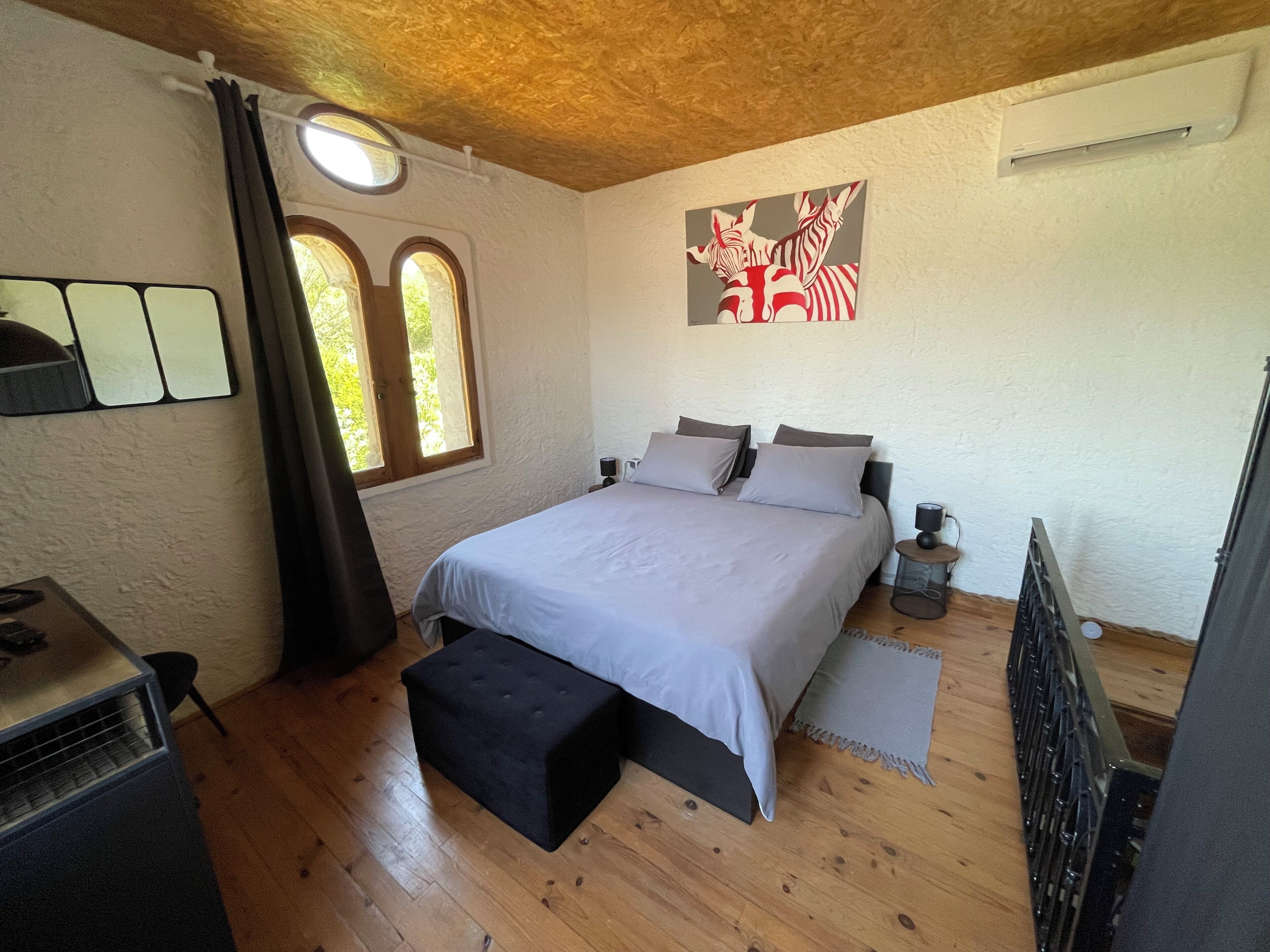 Un dormitorio con una cama, una mesita pequeña, un banco, un radiador y una ventana con cortinas.