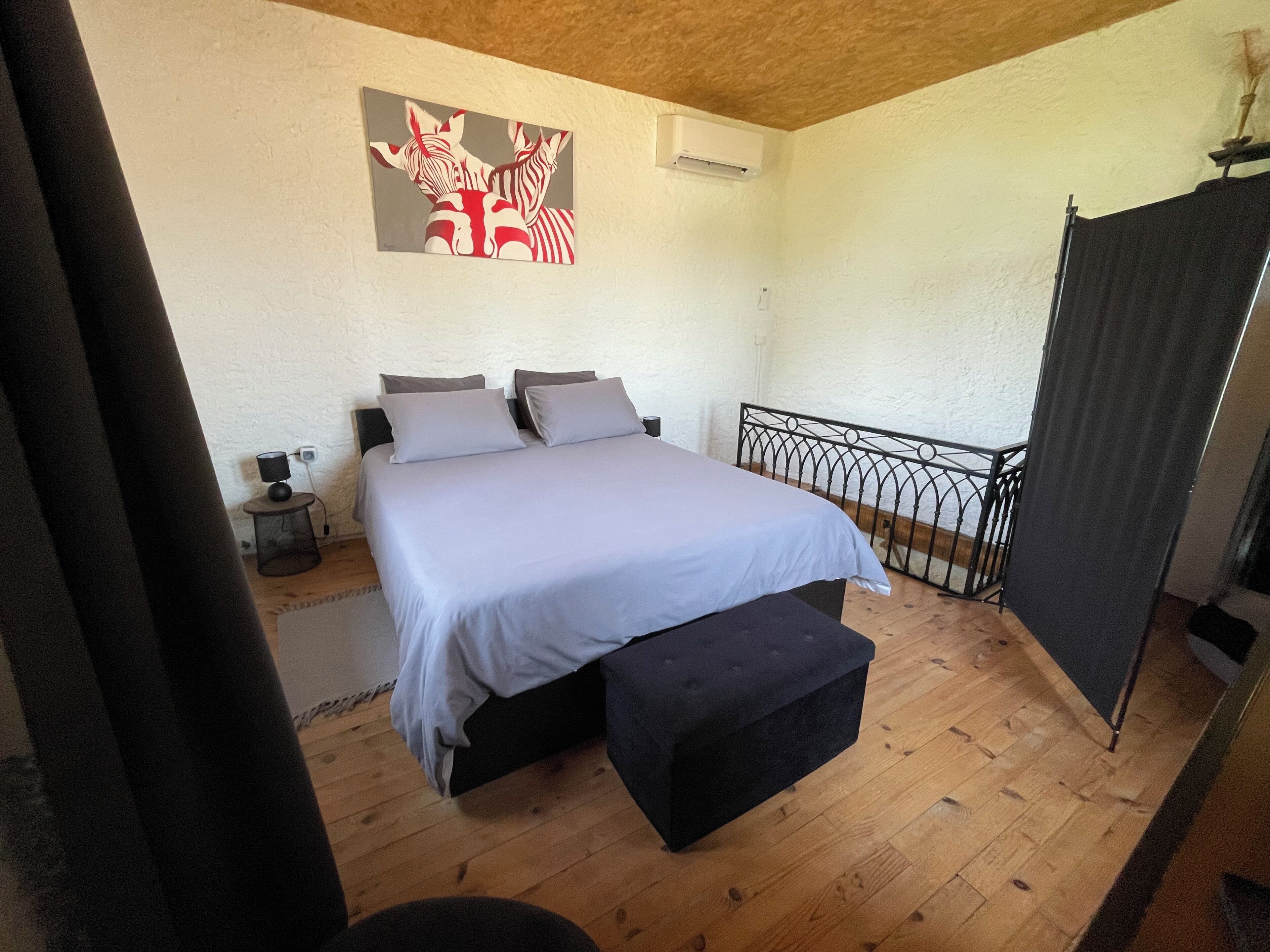 Une chambre à coucher comprenant un lit, des tables de chevet, un climatiseur fixé au mur, un banc noir et une rambarde en métal.