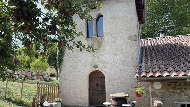 Un bâtiment rustique en pierre, doté d’une porte en bois, d’une petite table avec des tabourets et d’une plante suspendue.