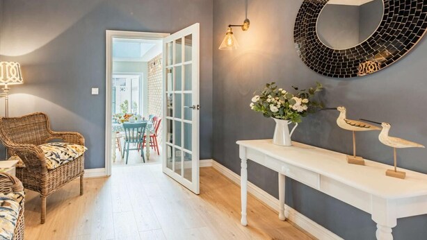 Una habitación con una silla de mimbre, una consola blanca, un espejo y una puerta que da a un comedor.