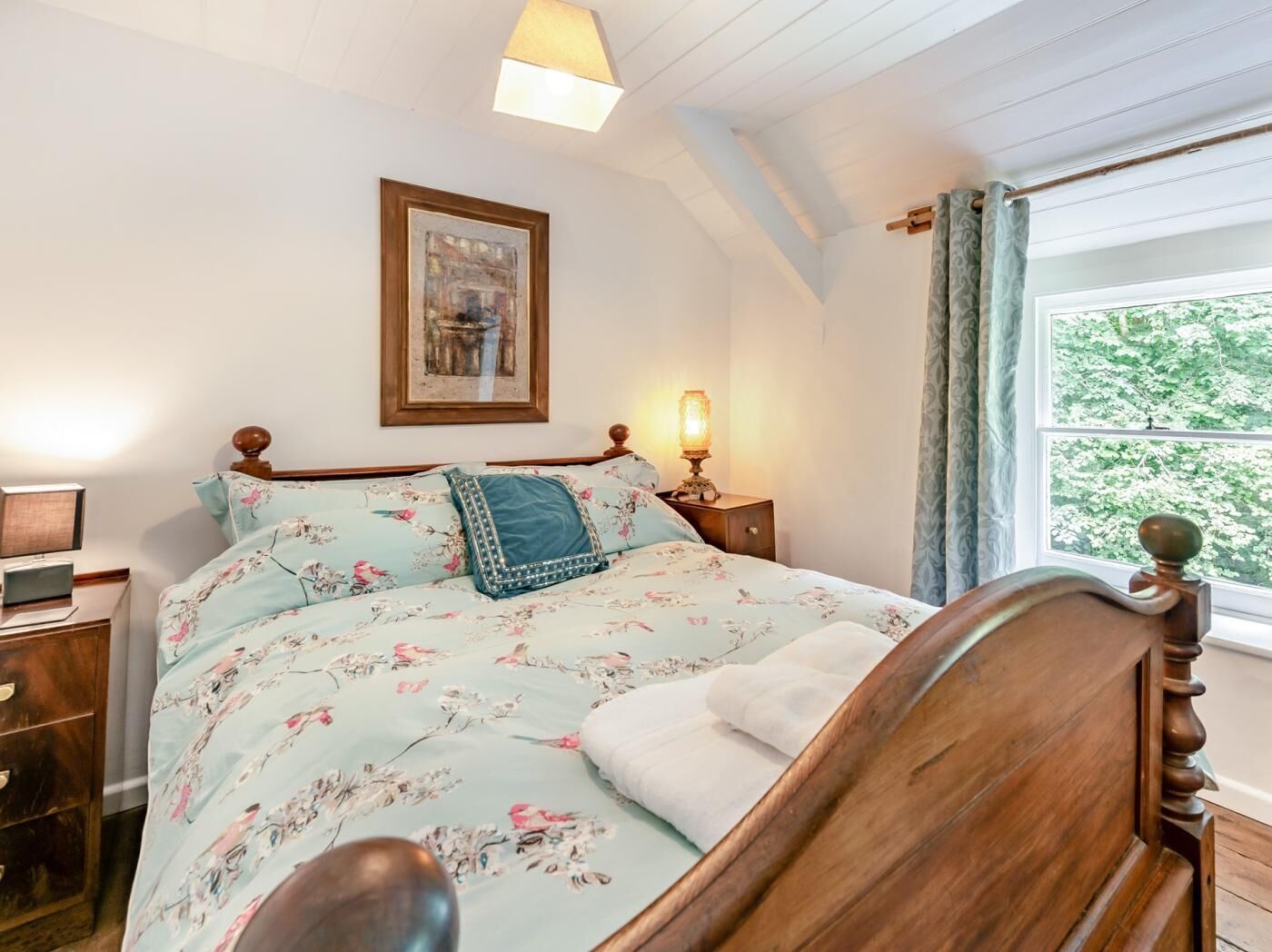 A bedroom with a wooden bed, a bedside table with a lamp, a framed picture on the wall, and a window with a view of greenery.