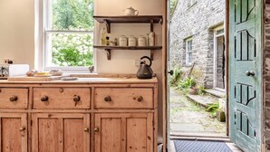 A rustic kitchen with wooden cabinets, a window with a view of greenery, and a stone wall outside. - 3 Bed in Lampeter (oc-55308) (Ciliau Aeron)