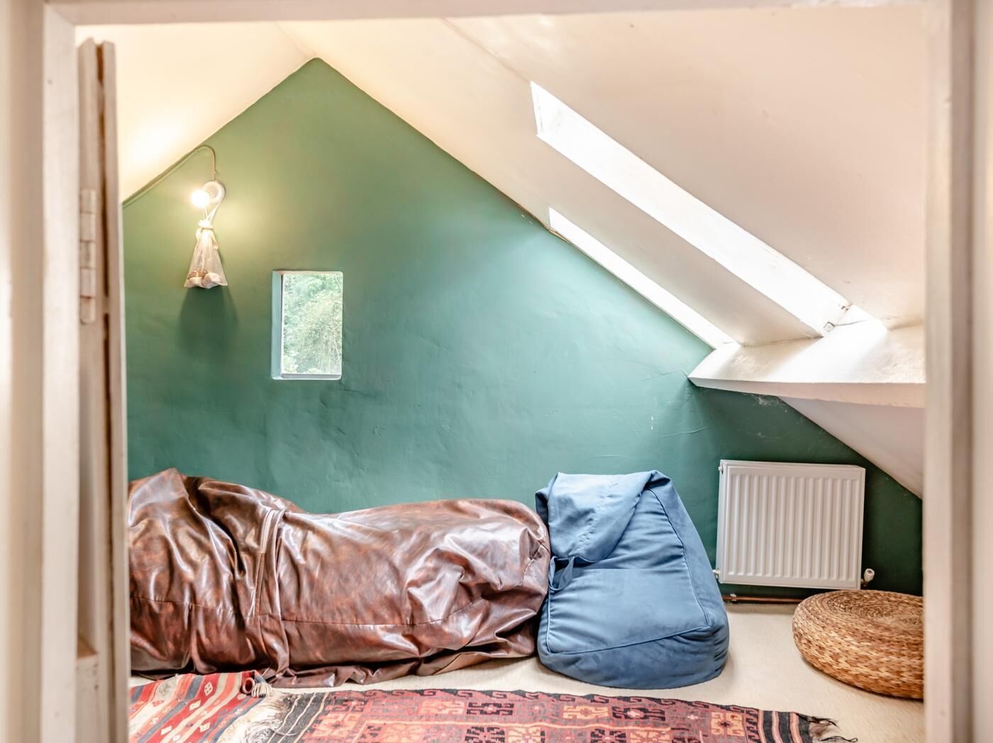 Una habitación con techo inclinado, un radiador, una cesta tejida, una alfombra estampada y una cama con cubrecama marrón y almohada azul.