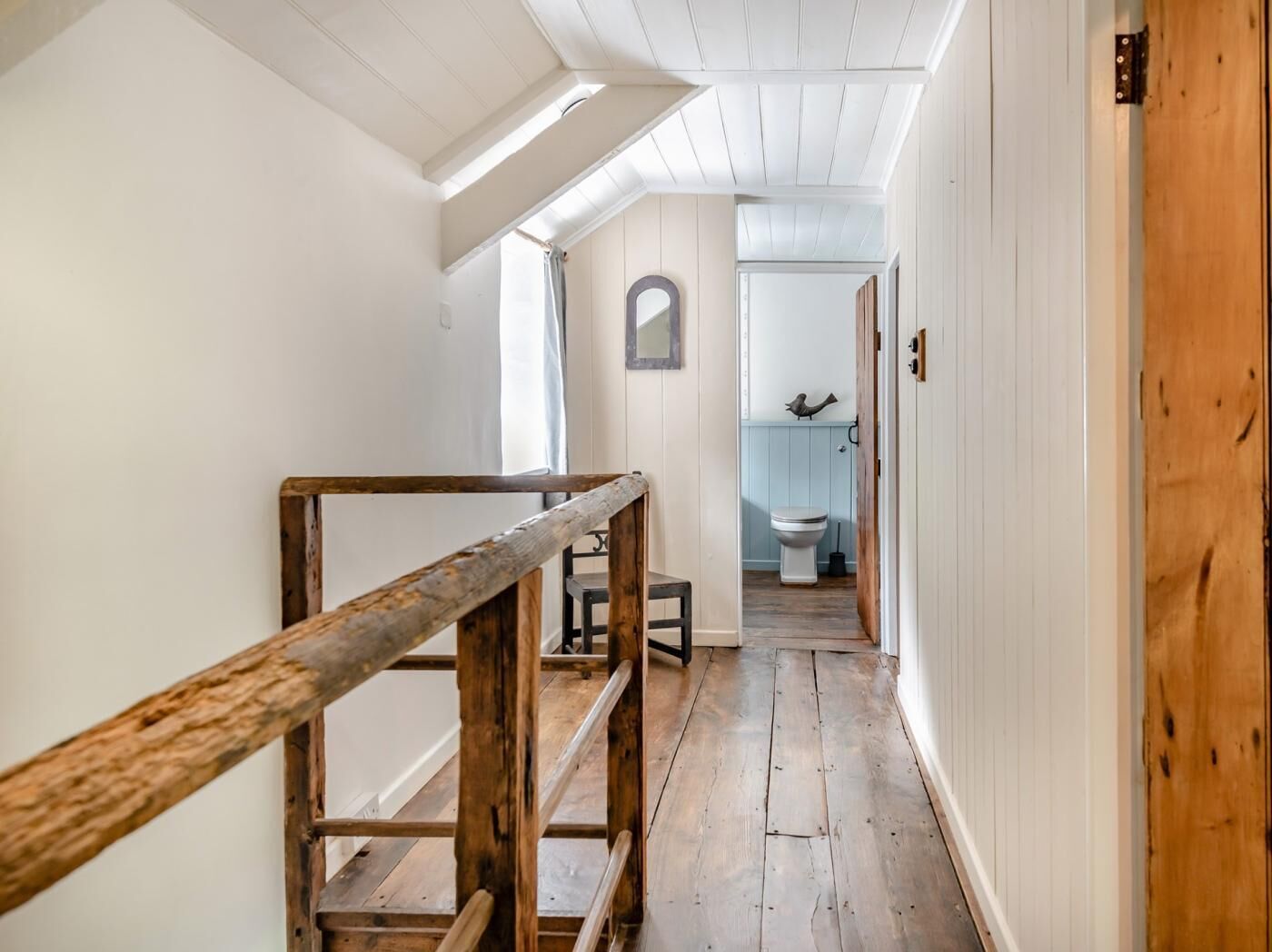 Un couloir étroit doté d’un plancher en bois, d’une rambarde en bois et offrant une vue sur une salle de bain avec des toilettes et une sculpture d’oiseau.