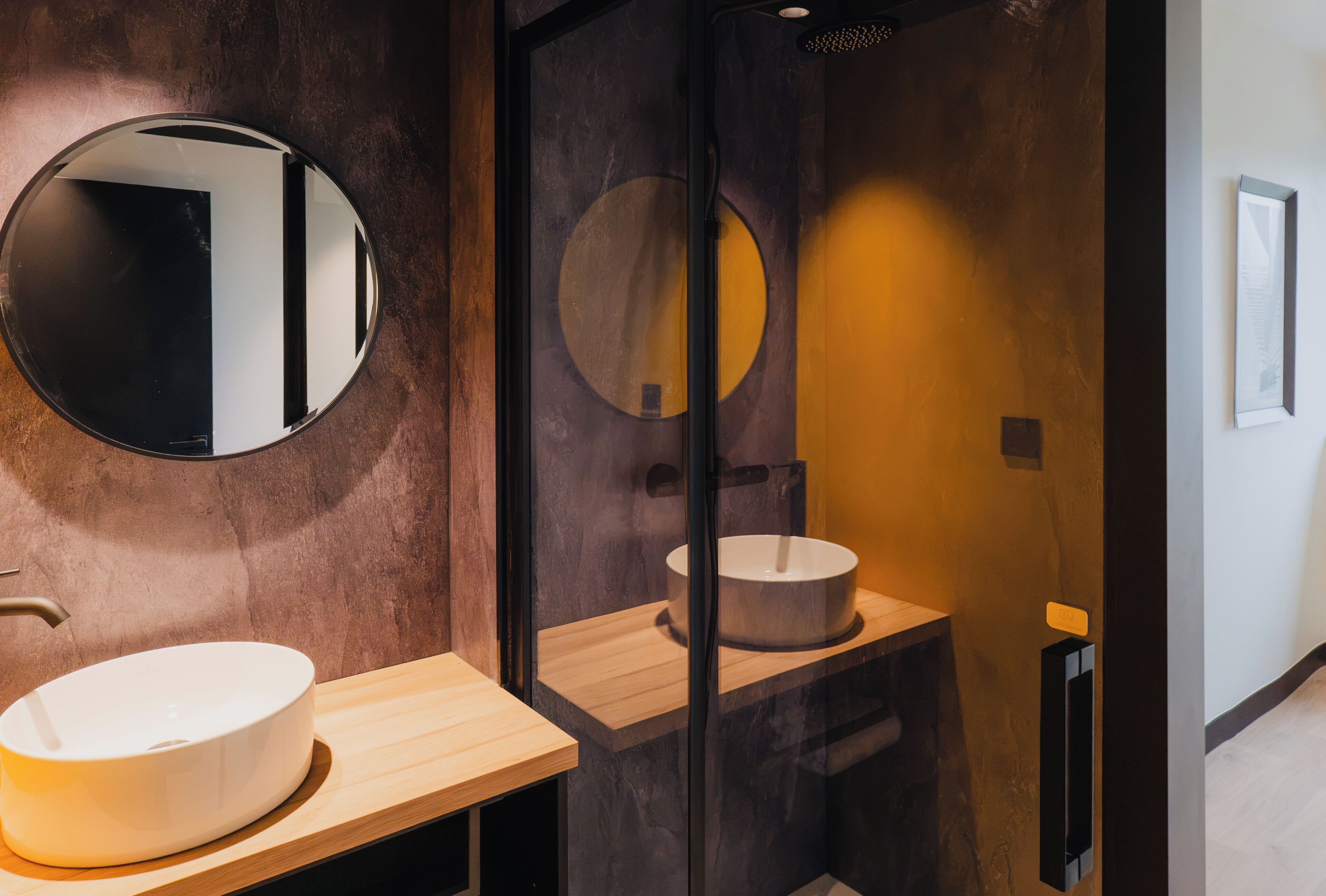 Une salle de bain moderne avec un grand miroir rond, un meuble-lavabo en bois et un lavabo encastré dans une structure en verre.