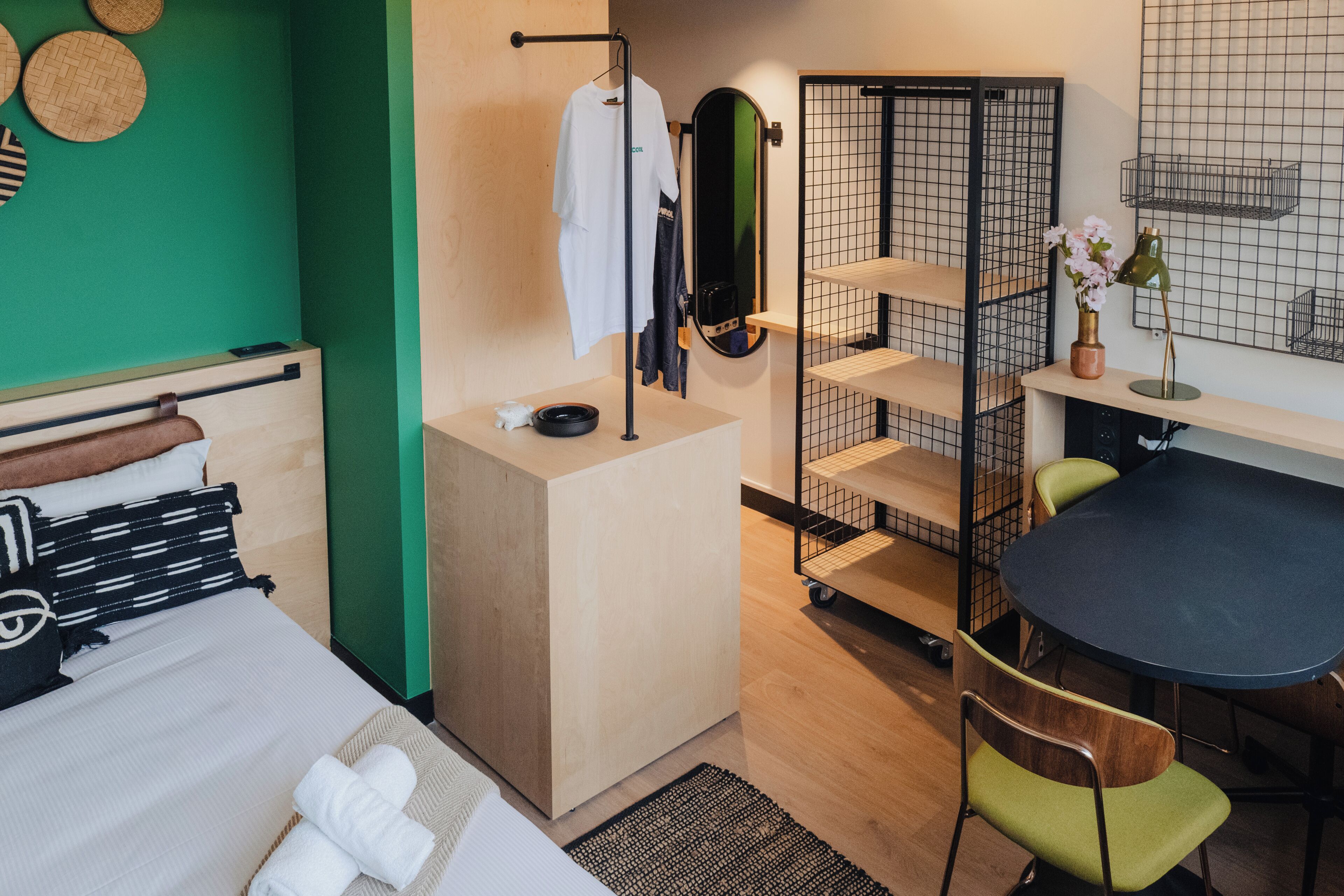 A modern hotel room with a bed, a desk, a chair, a mirror, and a small kitchenette.