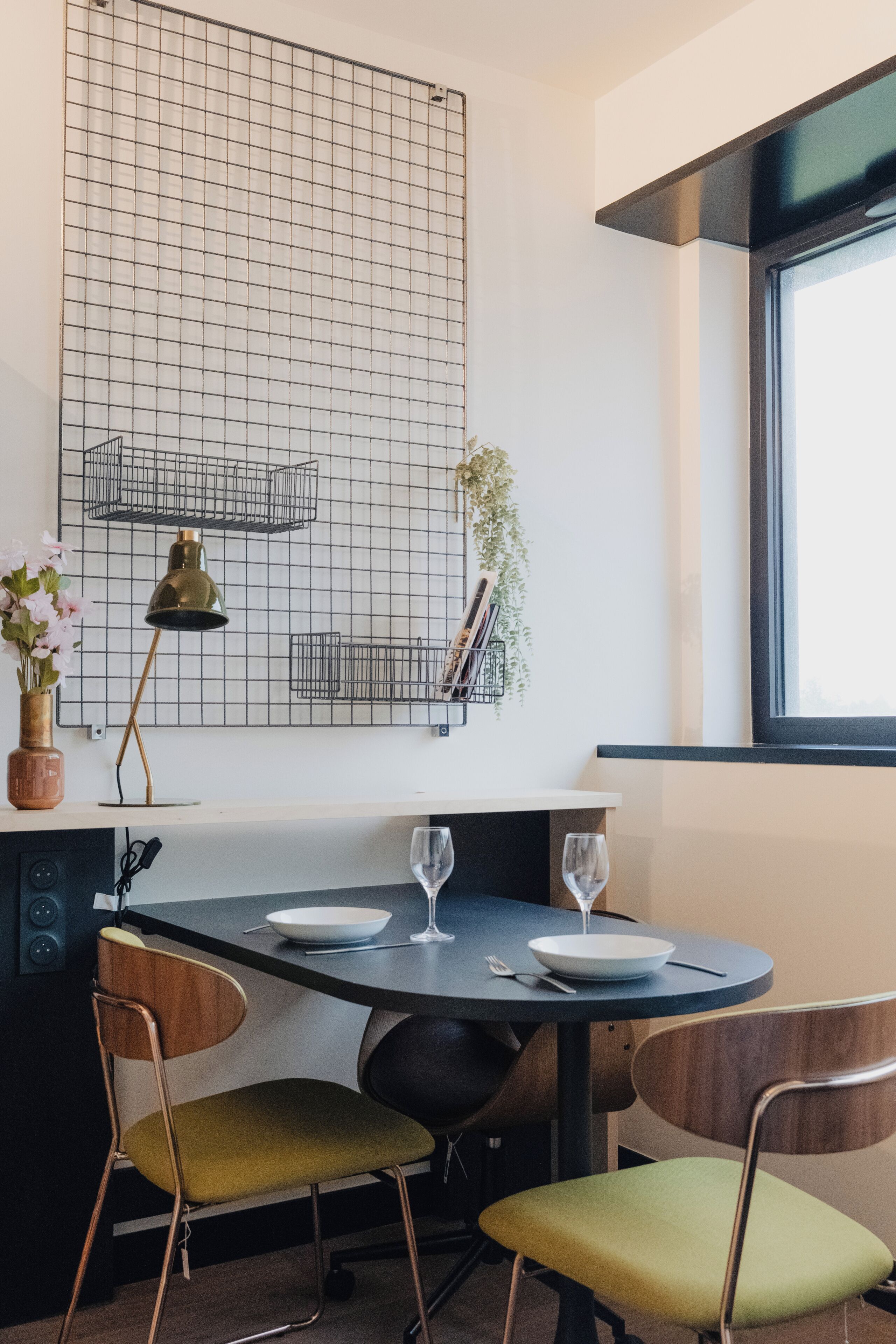 Een eethoek met een ronde tafel, twee stoelen, een vaas met bloemen, een lamp en een wand met een rasterpatroon.