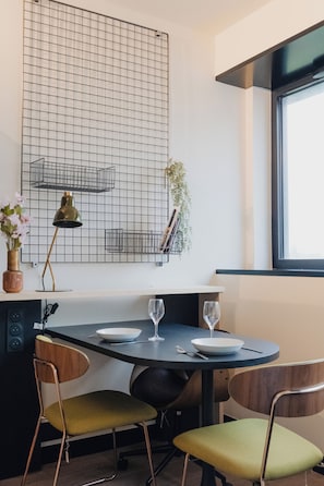 Un área de comedor con una mesa redonda, dos sillas, un florero con flores, una lámpara y un papel tapiz con diseño de cuadrícula.