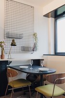 Un área de comedor con una mesa redonda, dos sillas, un florero con flores, una lámpara y un papel tapiz con diseño de cuadrícula.