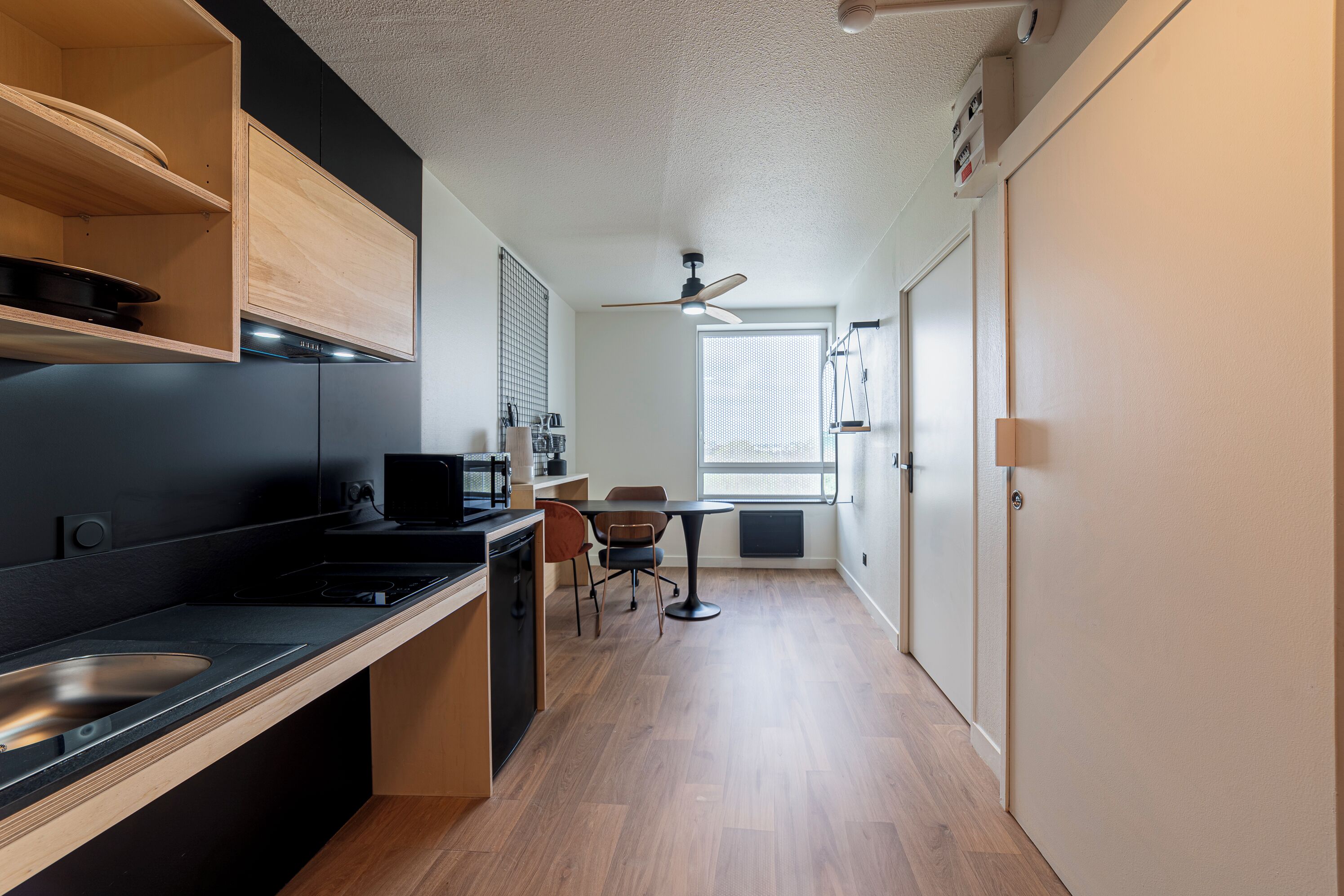 Una cocina compacta con microondas, estufa y lavaplatos. Hay un comedor con mesa y sillas. Se observa un ventilador de techo, y una ventana proporciona luz natural.