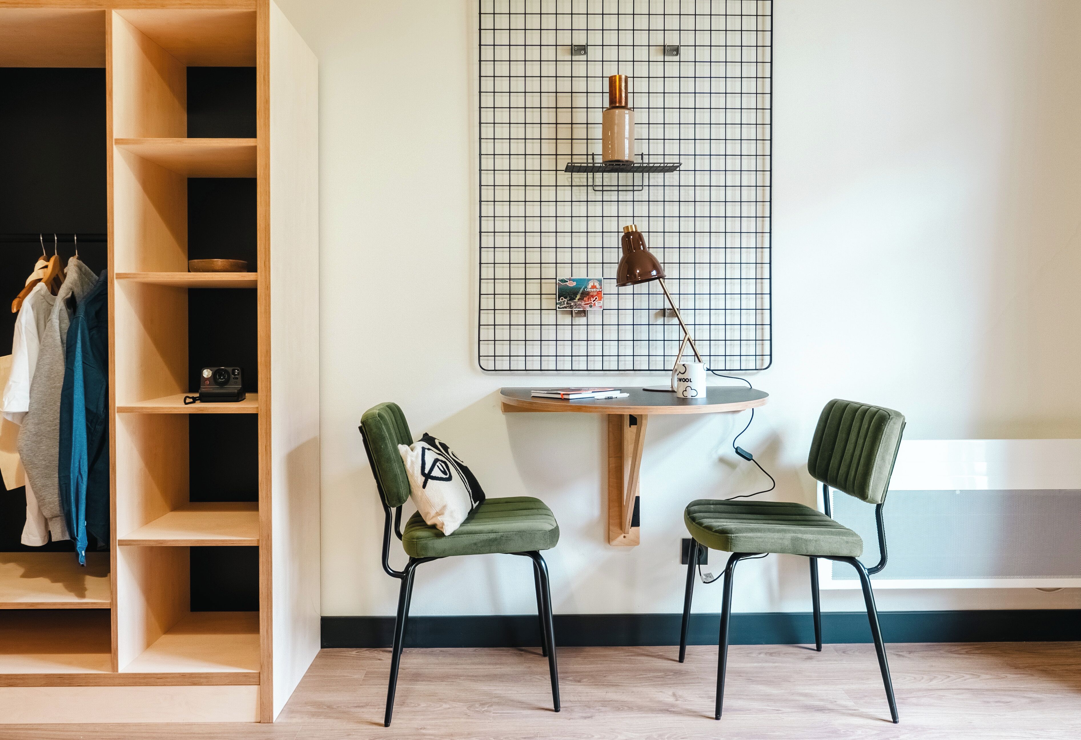 Una habitación con un armario de madera, dos sillas verdes, una mesita y una repisa en la pared con una lámpara y un marco de fotos.