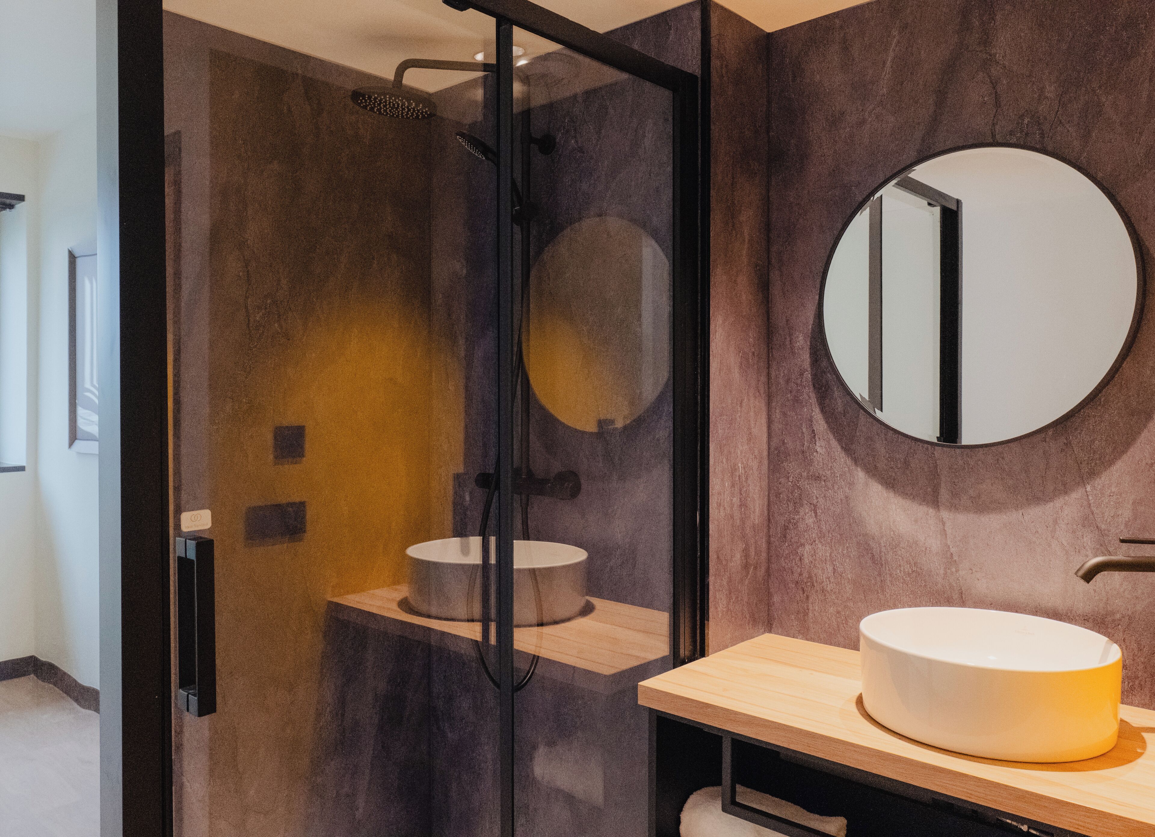 A modern bathroom with a glass-enclosed shower, a round mirror, and a wooden vanity with a sink.