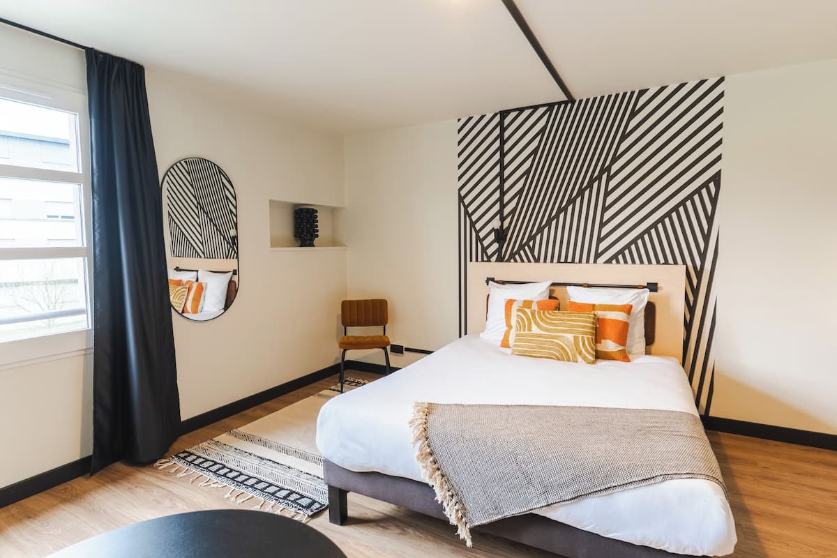 Quarto moderno com uma cama grande, mesa lateral, cadeira, espelho e cabeceira com padrão geométrico.