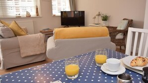 Ein Wohnzimmer mit einer Couch, einem Couchtisch mit einem Tischlein in Punkteoptik, einem Fernseher und einem Essbereich mit Essen und Getränken.