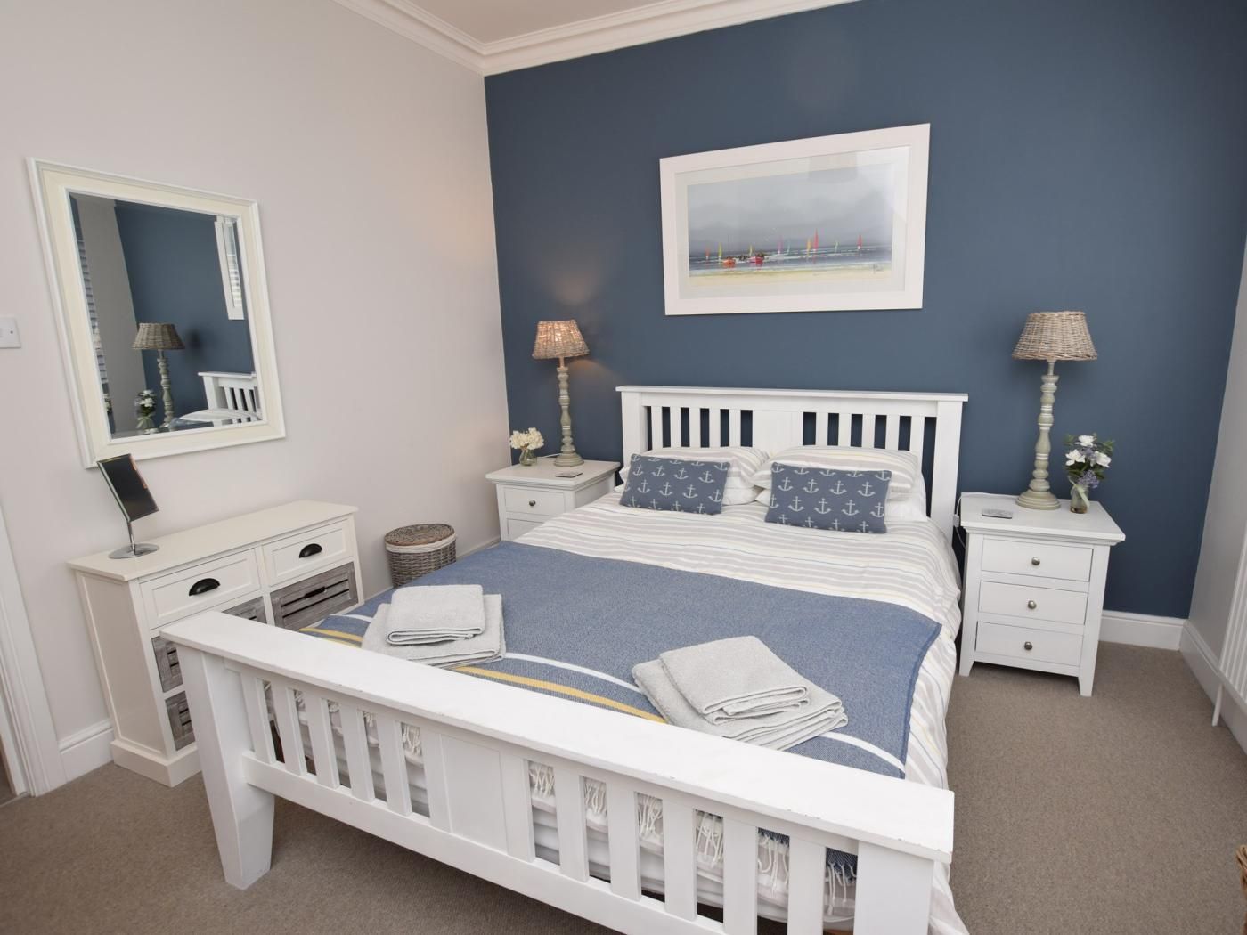 A bedroom with a white bed, a nightstand with a lamp, a dresser, and a mirror.
