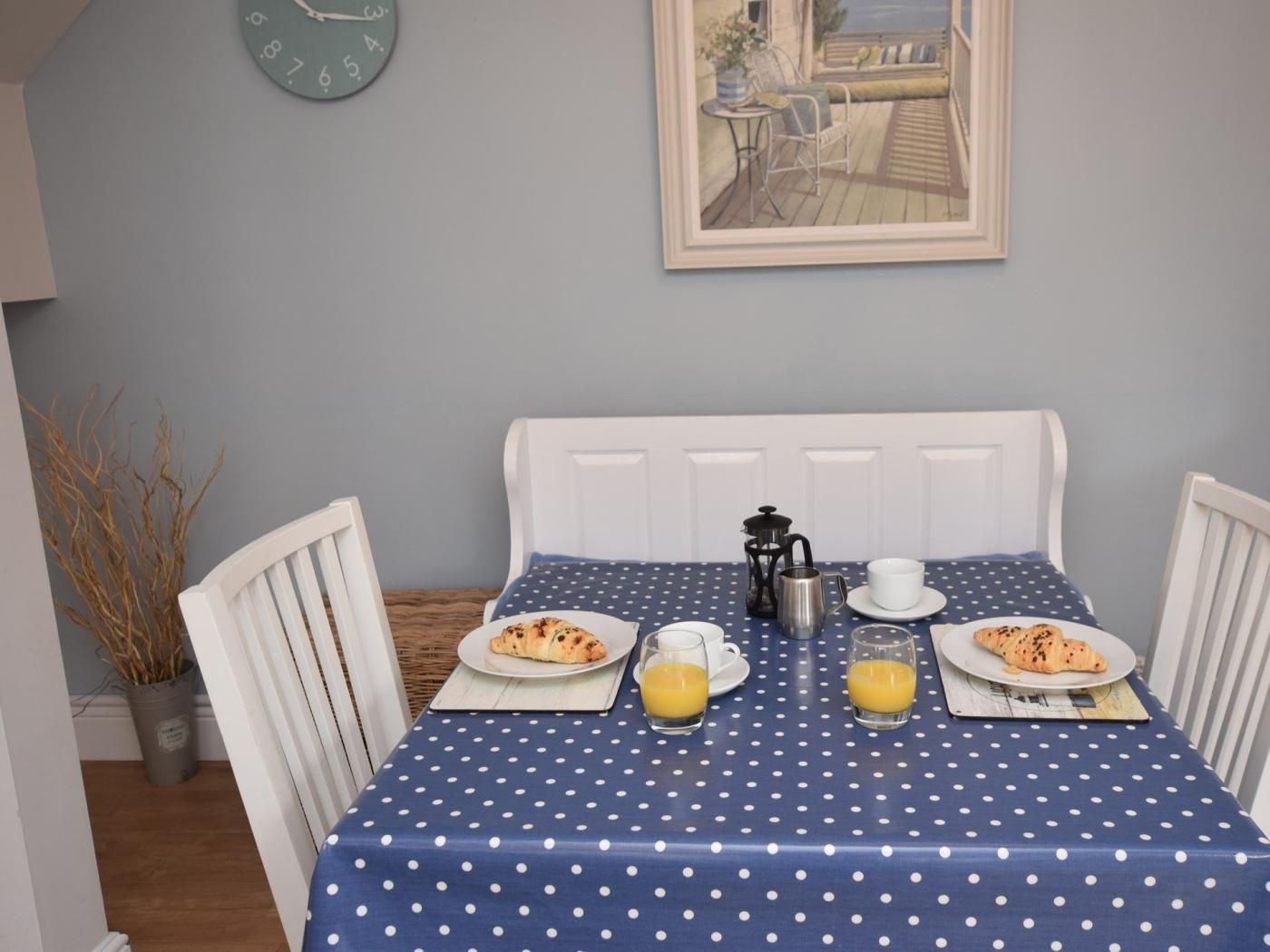A dining table with a polka dot tablecloth, two plates of food, two glasses of orange juice, a coffee pot, and a clock on the wall.