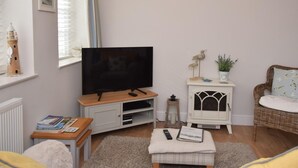Ein Wohnzimmer mit Fernseher, einem kleinen Tisch mit Büchern, einem Korbsessel, einer weißen Heizung und einem Fenster mit Vorhängen.