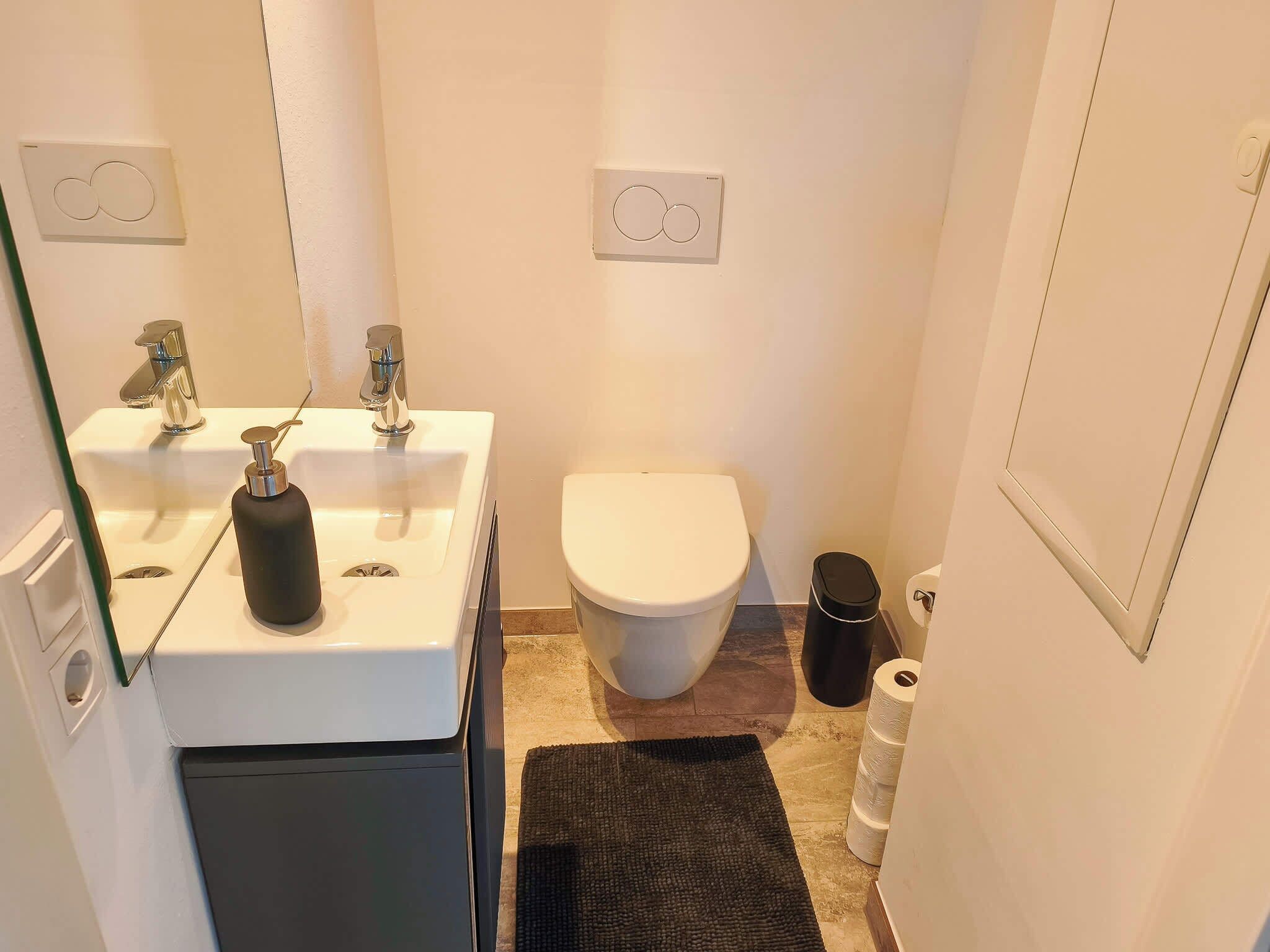 Un baño pequeño con un lavamanos blanco, dos grifos, un inodoro, un cubo de basura negro y un rollo de papel higiénico.