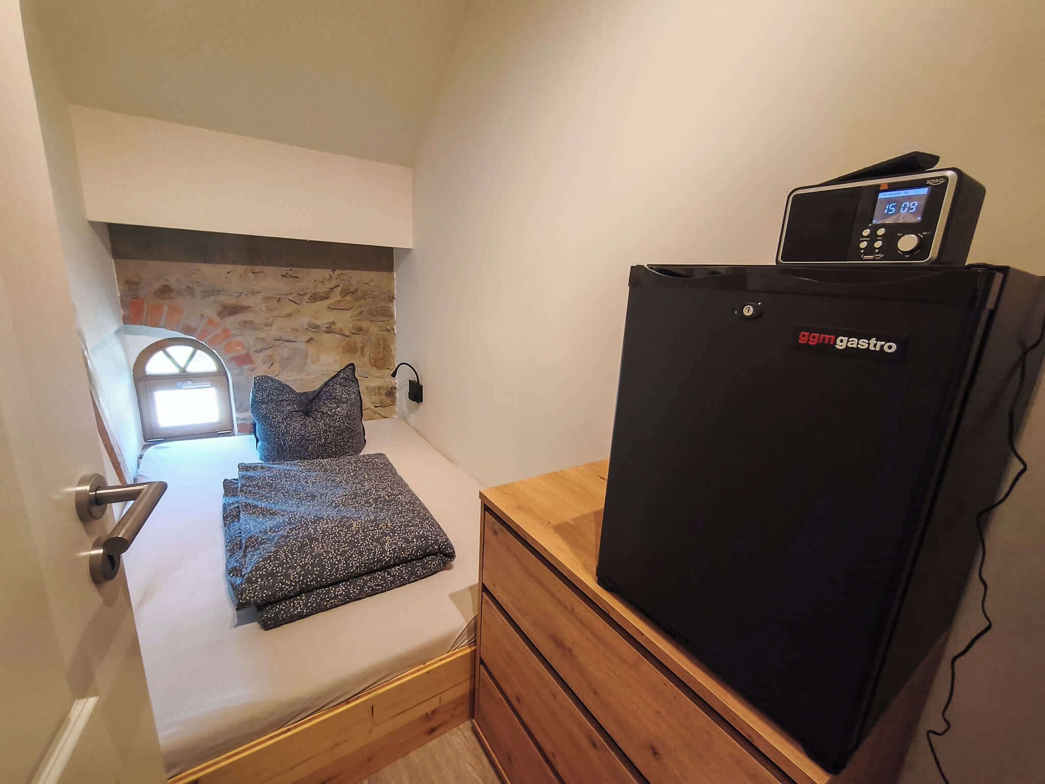 Una habitación pequeña con una cama, un aparador de madera y un refrigerador negro con pantalla digital.