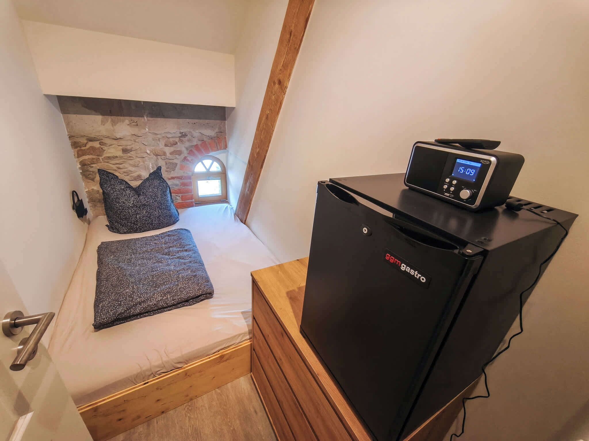 A compact room with a bed, a small black refrigerator, and a digital clock.