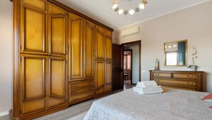 Quarto com um guarda-roupa de madeira grande, uma cama com colcha branca, cômoda e um espelho na parede.