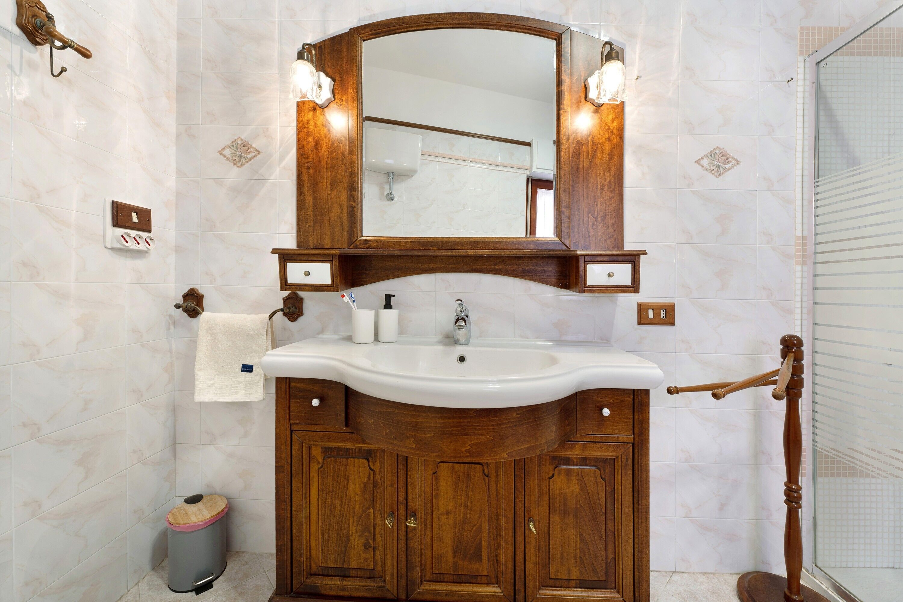 A bathroom with a wooden vanity, a sink, a mirror with lights, a towel rack, and a trash bin.