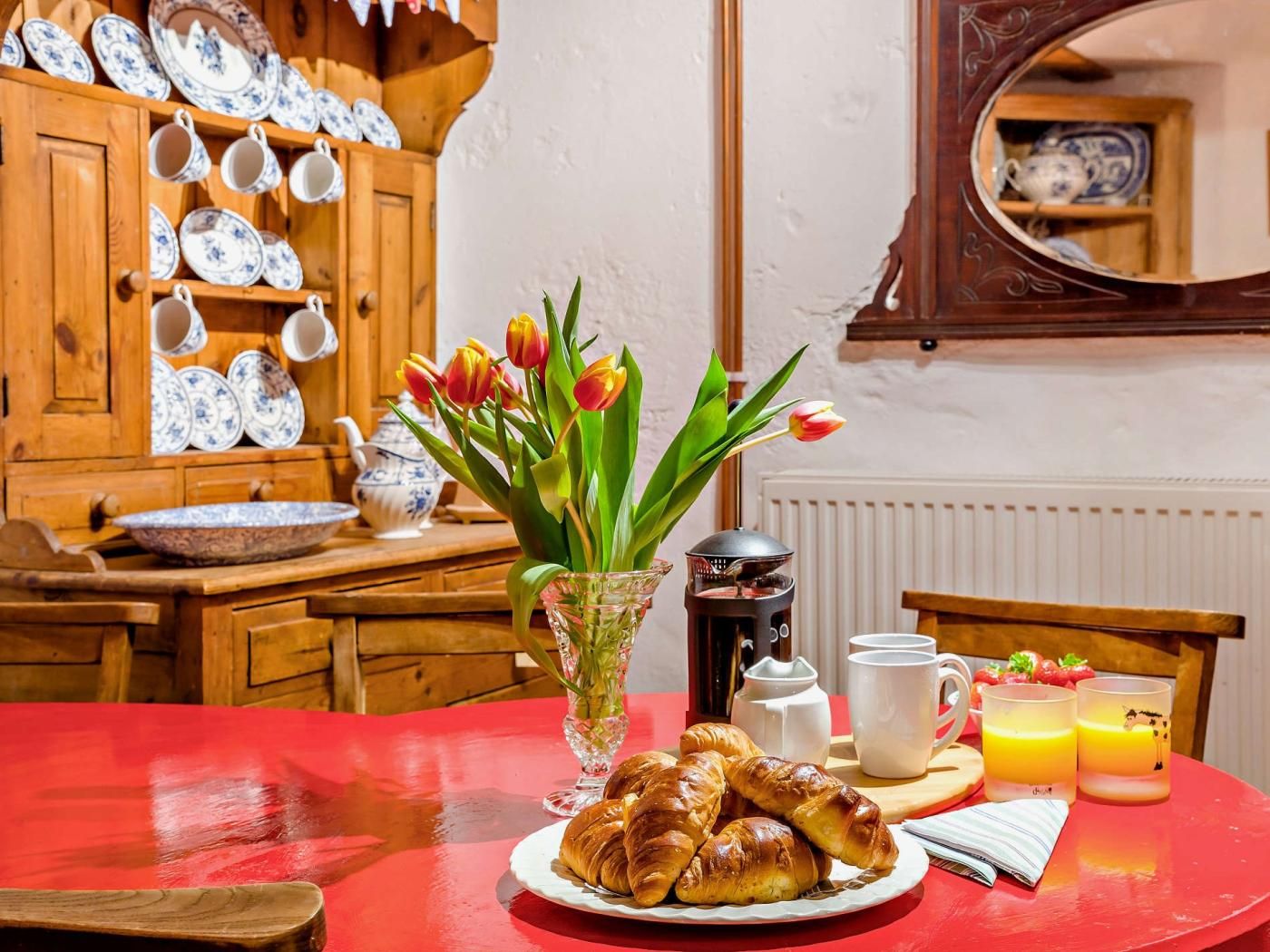 Un desayuno con una mesa roja, un plato de cruasanes, un jarrón de tulipanes y una cafetera.