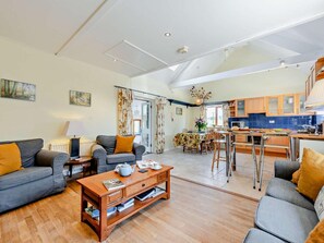 Ett rymligt vardagsrum med en soffa, ett soffbord och ett matbord med stolar. Ett kök med träsk åp och en matplats med en kristallkrona syns i bakgrunden.