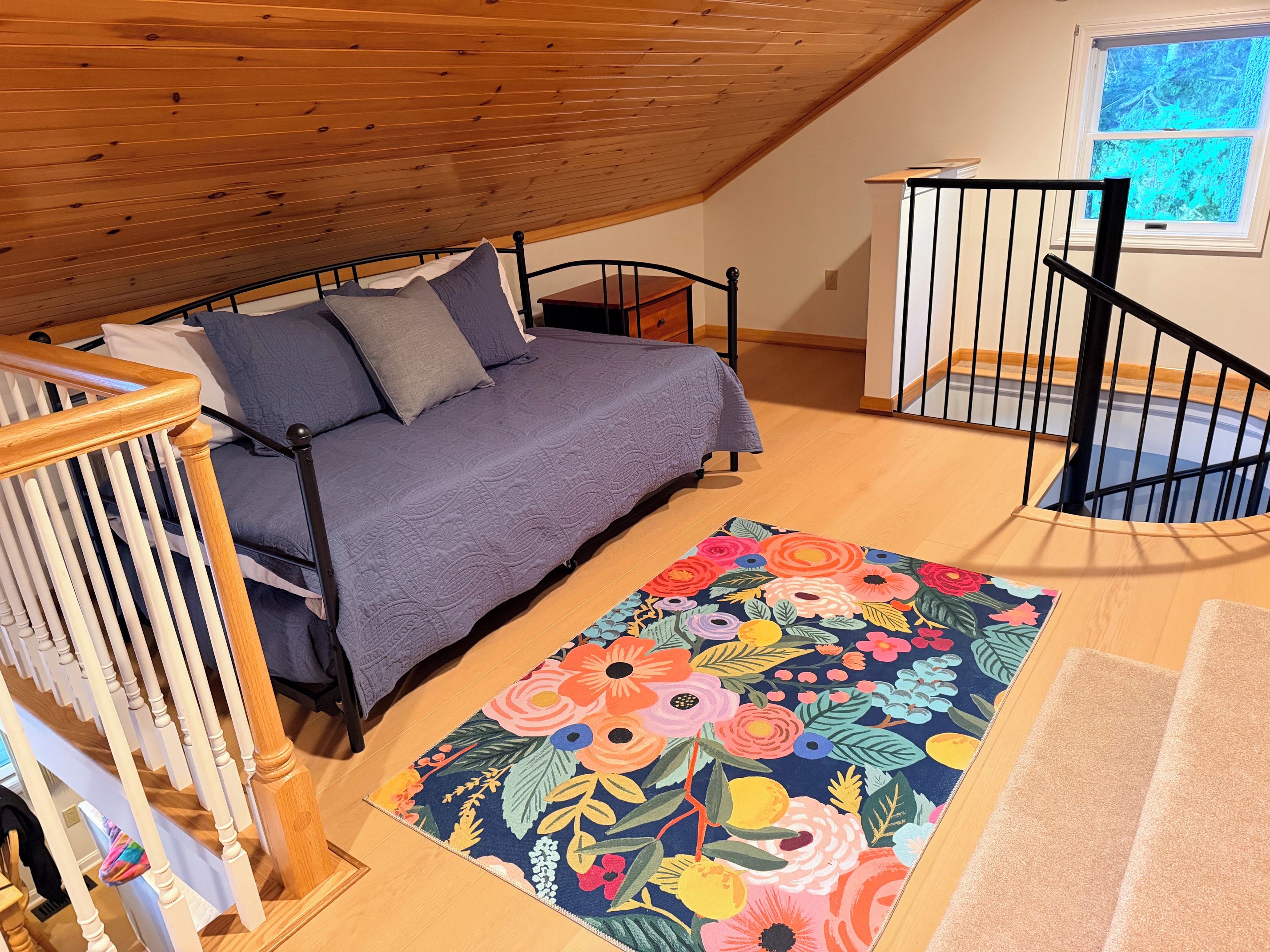 A bedroom with a bed, a wooden ceiling, a window, a staircase, and a colorful rug.