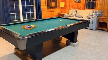 Una mesa de billar en una habitación con paredes de madera, un sofá con cojines y una lámpara.