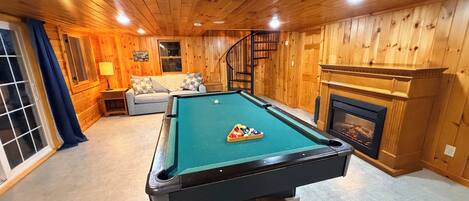 A pool table in a room with wooden walls, a sofa, and a fireplace.