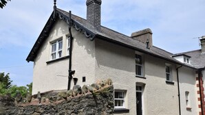 A two-story building with a stone wall in front, a chimney, and a window with a door. - 2 Bed in Penmaenmawr (oc-56081) (Penmaenmawr)