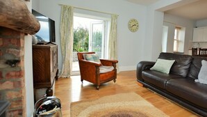 A living room with a fireplace, a wooden cabinet, a leather sofa, and a large window with curtains. - 2 Bed in Penmaenmawr (oc-56081) (Penmaenmawr)